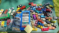 Overview of assortment of about 40+ toy cars including loose cars and some packaged sets spread on a green surface outdoors