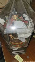 Music box with Christmas holiday figurines, miniature trees, candles, and gifts inside a multi-faceted glass display case with a dark base, viewed from one angle.