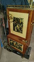 Front angled view of vintage wooden rolling bar cart showing painted panels with artwork, brass wheels, and side handle.