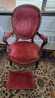 Antique armchair upholstered in deep red velveteen fabric with carved wooden frame, claw feet on casters, and matching red velveteen footstool in front