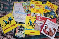 Photo of all 13 children's and cartoon books spread on patterned carpet, shows title and colorful illustrated covers including several Curious George books and others.