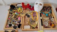 Overall view of the costume jewelry lot showing various colorful jewelry pieces, a white decorative mask, cat-faced wristwatch, and sunglasses spread on three wooden trays.