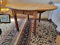 Angle view of the oval oak dining table showing the drop leaf extended and four turned legs, placed over patterned rugs.