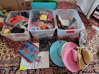 Wide view of three large plastic bins filled with packaged silk scarves and embroidered jewelry bags, plus pillow cases featuring bright animal prints, and four colorful straw sun hats stacked on floor