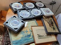 Top down view of 6 Limoges collector plates in foam holders on a wooden table alongside framed prints with maritime and equestrian scenes.