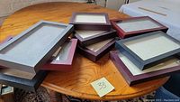 Ten rectangular ring display boxes stacked on a wooden table. Various colors visible including black, gray, burgundy, and dark blue. Each box has a transparent glass lid showing the interior lined with soft fabric and slots designed to hold rings.