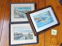 Three framed watercolor paintings laid on wooden floor, showcasing seaport scenes.