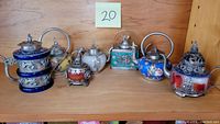 Seven small wine pots lined on wooden shelf, varied shapes, enamels and silver mounts.