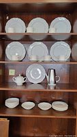 Shelf showing multiple stacked and arranged white Federalist Ironstone plates, cups, and saucers, with a sugar bowl and metal creamer pitcher, placed against a wooden cabinet backdrop.