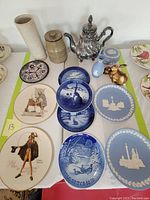 Photo of the overall lot containing collectors plates, pottery, and a silver teapot on a table with items arranged flatly.