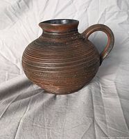 Side view of the round, ribbed pottery vase with handle on a white fabric background.
