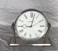Front view of Howard Miller desk mantel clock showing white clock face with Roman numerals and silver plated frame with supporting rods and base