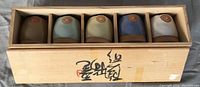 Wooden box holding five Japanese ceramic tea cups with slots and calligraphy on the top.