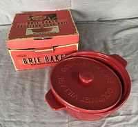 Red ceramic brie baker with embossed lid, showing the brand Gourmet Village, positioned next to original red box indicating it is new in box.