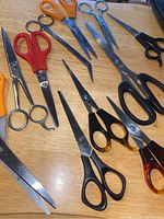 Photo showing an assortment of eleven scissors on a wooden surface, displaying different sizes, shapes and handle colors including orange, red, black, and blue.