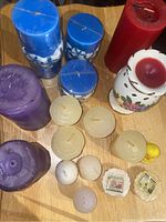 Photo showing assorted candles in various colors and sizes arranged on a wooden surface, including blue, red, purple, beige, ball shaped, chick shaped candles and the ceramic holder with floral design.