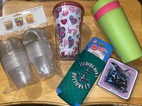 Photo showing clear 5.5 oz cups in original plastic packaging, a tumbler with hearts and butterflies design with lid, a green tumbler, green can cooler with anchor logo, and set of SeaWorld coasters.