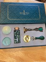 Full view of wax sealing kit box with two tealight candles, glass vial of green wax beads, melting spoon, and metal stamp