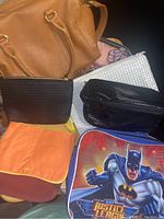 Photo showing a close-up of nine assorted bags and wallets grouped together on a dark surface. Includes tan handbag, black wallet, orange fabric purse, white clutch with texture, and Batman Justice League pouch with comic graphic.