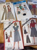 Photo shows all five sewing pattern envelopes arranged on a wooden surface. Patterns include women’s dresses, skirts, shorts, and pants in various styles. The Simplicity brand and pattern numbers are clearly visible.