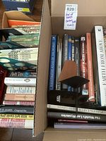 Box containing a mix of paperbacks, some hardcover coffee table books, and two wooden bookends inside a cardboard box.