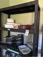 Photo showing black plastic shelving unit with three levels of cookware and stoneware items, including The Rock cookware, Buchan stoneware, and other baking dishes.