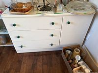 Front view of modern white dresser with green knobs, showing three drawers and a door with contents on top not included.