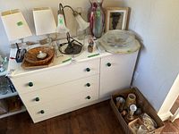 Photo of white dresser top holding two clear crystal base lamps with white square shades, a 17 inch flower-patterned lamp without shade, a tall sparkly glass vase, framed 3D grape art, glass platters, and assorted small decorative items. A box of battery-operated candles and other household items are to the side.
