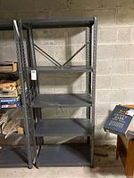 Front view of the full metal shelving unit, showing five shelves and overall tall rectangular form, placed against a concrete block wall.