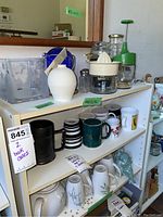 Photo shows shelves with kitchen and decorative items including Corelle tea pot and mugs, various coffee mugs, foos choppers, salt and pepper shakers, and angel figurines.