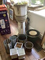 Overview photo showing a tall cream ceramic pedestal with live plant on top, surrounded by various pots in ceramic and metal including glazed pots, white square ceramic pots, and metal bucket-style pots.