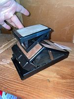 Folded Polaroid SX-70 Land Camera Model 3 resting on a wooden surface, showing the top and side with worn leather surface and black plastic.