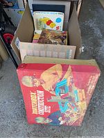 Photo showing a large vintage Matchbox Construction Site kit box, red in color with children playing with toy construction vehicles on the front.