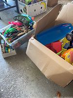 Wide shot of cardboard box and clear plastic bin filled with assorted plastic figurines, toy cars and trucks, and toy planes.