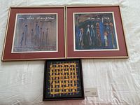 Three framed artworks laid flat on a white surface. Two larger pieces have abstract figures and French text with pink mats and gold frames. One smaller piece has a dark frame with yellow figure weaving pattern on black background.
