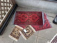Two small square brown decorative carpets placed on top of one of the two large red and black patterned carpets on a concrete floor.