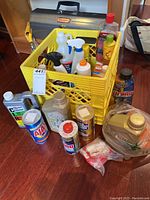 Photo showing crate filled with assorted cleaning sprays and bottles inside, surrounded by several additional cleaning product bottles and cans.