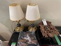 Overview of two brass table lamps with cream lampshades alongside wooden cuckoo clock parts on table