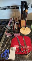 Photo showing multiple large kitchen knives with black handles, various scissors, and a wooden knife stand containing knives.