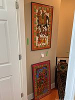 Two framed painted cloth artworks, one hung vertically on the wall and the other leaning against the wall, both showing colorful traditional scenes with human figures and an elephant.