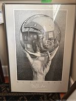 Framed M.C. Escher poster showing hand holding reflective sphere with room interior and figure reflected, signed M.C. Escher
