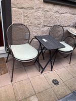 Photo of two brown metal mesh garden chairs with beige cushions and a small black folding wooden table between them, set on an outdoor paved area.