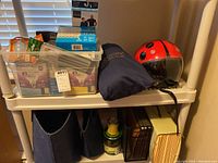 Shelf with red ladybug helmet, blue travel pillow in pouch, plastic tub of light bulbs, canned goods, and wire file organizer with folders.