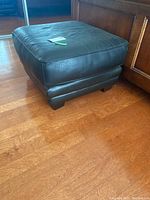 Black faux leather square ottoman placed on wooden floor with cushioned top and block base.