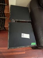 Top and side view of two square black side tables placed on wooden floor next to brown leather furniture.