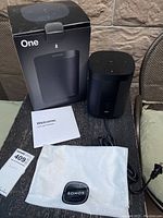 Sonos One speaker with original packaging, power cable, instruction booklet, and cloth bag placed on a black table against a stone wall background.