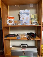 Wooden shelving unit containing a boxed VTech cordless phone, white coiled coaxial cable, assorted phone hardware, black handheld power tool, black and red handheld device, and silver Craftsman toolbox.