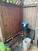Four red metal trellises leaned against a wooden fence with black metal stands, a blue plastic bin with lid, and a white bin with gardening tools.
