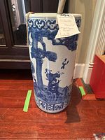 Front view of the umbrella stand showing Chinese figures and tree scene in blue on white ceramic with attached tag.