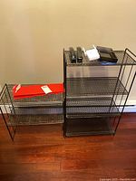 Photo of two black metal wire shelving units side by side on a wooden floor against a wall, with several items on top including a TP-Link router and handheld devices.